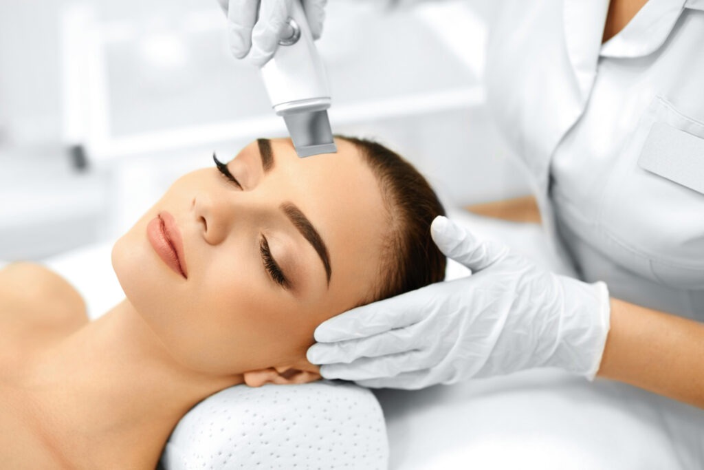 A woman receiving a facial treatment, with a beautician using a skincare device on her forehead. The serene setting emphasizes relaxation and skincare.