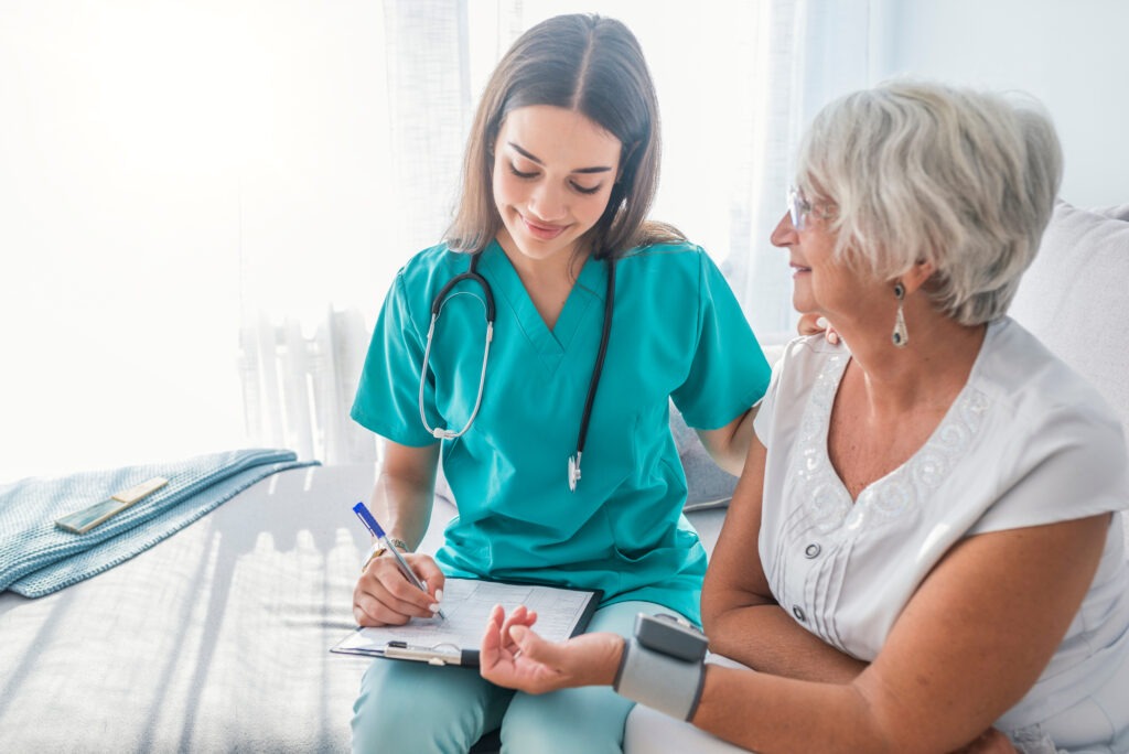 A healthcare professional in scrubs records notes while checking the blood pressure of an elderly woman in a bright, comfortable clinic setting.