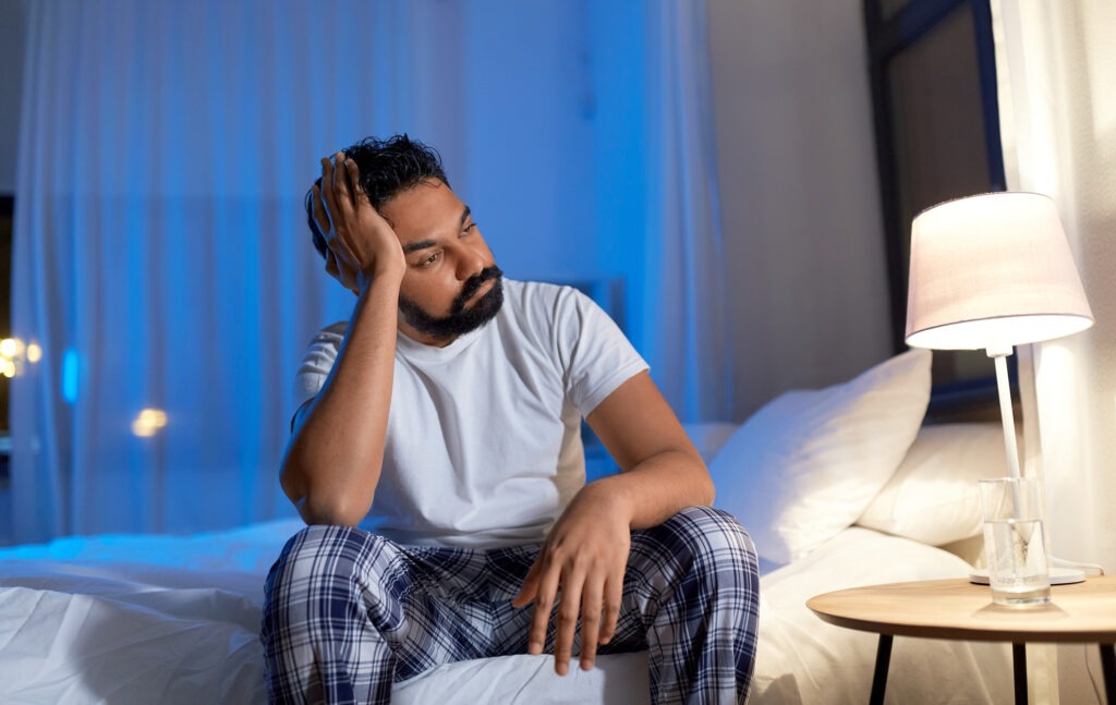 A man sits on a bed, looking contemplative and stressed, in a softly lit room with blue tones, conveying feelings of anxiety or sleeplessness.