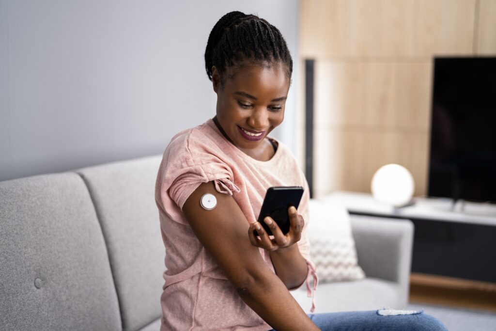 A young woman smiles at her smartphone while monitoring a wearable health device attached to her arm, showcasing health technology in everyday life.