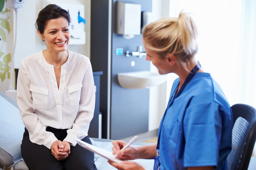 A smiling patient in a white blouse engages in conversation with a nurse in blue scrubs, discussing health information in a medical office.
